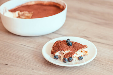 A piece of tiramisu with cocoa, blueberries and tender cream on a plate on a wooden table.