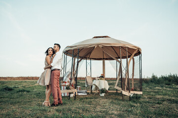 Full length portrait of young couple dating near trasparent tent with served table. Newlyweds celebrate their honeymoon and spend time together outdoors. Romantic dinner concept. Image with copy space