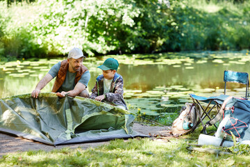 Full length portrait of father and son setting up tent together while enjoying camping by lake in forest, copy space
