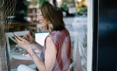 Rear view of female blogger using mobile phone for checking mail and messages notification on break, smiling beautiful woman holding smartphone sending content and and text in chat app at cafe