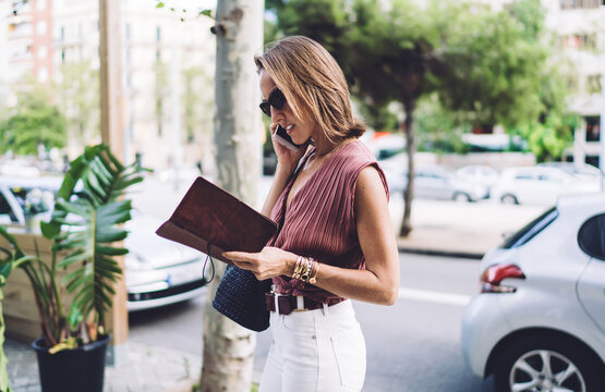 Stylish adult woman with notebook speaking on smartphone on street
