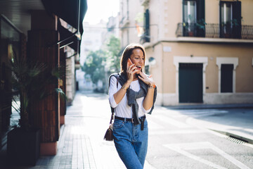 Fototapeta premium Smiling caucasian woman in casual wear walking on city street talking on mobile phone on sunny day, positive young female making smartphone conversation walking in town making call in roaming