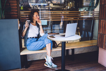 Pensive caucasian female in casual wear sitting on cafe terrace with technology on coffee break, serious businesswoman drinking beverage while working on laptop computer on freelance job process