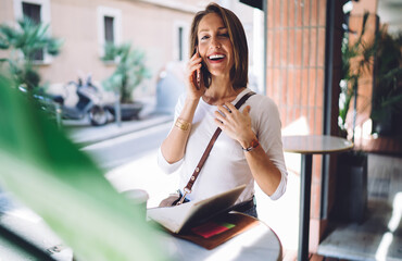 Happy caucasian woman enjoying conversation on mobile phone resting in cafeteria on free time, smiling female millennial satisfied with tariffs and connection on cellular having positive conversation