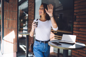 Woman in casual outfit with coffee smiling and waving hand
