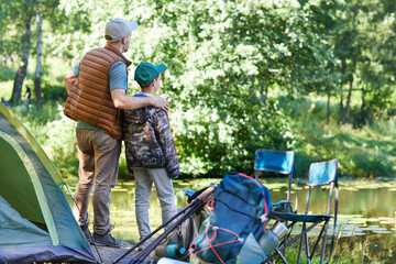 Back view full length portrait of father and son looking at lake while enjoying camping trip together, copy space