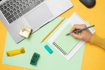 Workspace with a laptop, the hands of a girl writes a list of gifts in a notebook, a gift box and 25 on a yellow background. Layout of the apartment, ordering gifts online, top view.