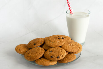 Milk in a glass with a straw, next to oatmeal cookies with chocolate chips. Christmas drinks. White marble background. Selective focus. Copy space. Banner