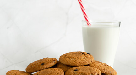 Milk in a glass with a straw, next to oatmeal cookies with chocolate chips close-up. Christmas drinks. White marble background. Selective focus. Copy space. Banner