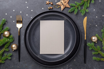 Christmas table setting. Black ceramic plate with blank sheet for your text, fir tree branch and accessories on stone background. Gold decoration