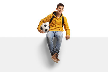 Teenager boy holding a soccer ball and sitting on a white panel board