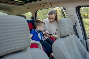 Fototapeta premium Mother gives the phone little boy sitting in a car seat. Safety of transportation of children.