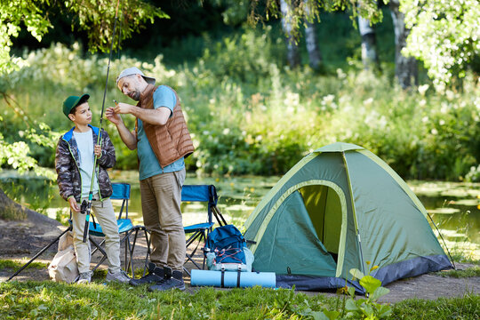 Full length portrait of loving father teaching son to set up fishing equipment while enjoying camping trip together, copy space