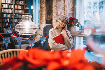 Portrait of beautiful hipster girl in trendy cap holding personal diary and looking at camera during rest time in loft cafeteria with X-mas decoration, Caucasian woman with education textbook posing