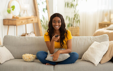 Fototapeta premium Home entertainments. Smiling black woman with remote control eating popcorn and watching TV on sofa in living room