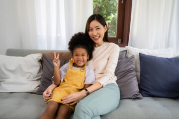 Blured defocused A happy Asian mother hugging her African daughter who made a peace sign hand gesture sitting together on sofar with many pillows in a living room, they both have a big smile.