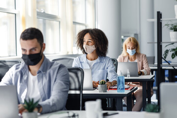 Generate an idea and inspiration for work during covid-19. Focus on african american woman in protective mask looks out the window at workplace with laptop and antiseptic