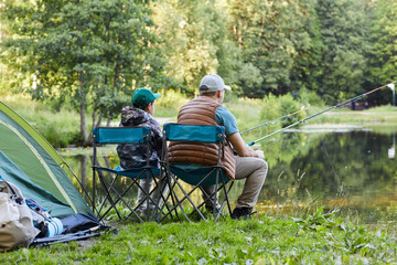 Back view portrait of loving father and son fishing by lake together during camping trip in nature,...