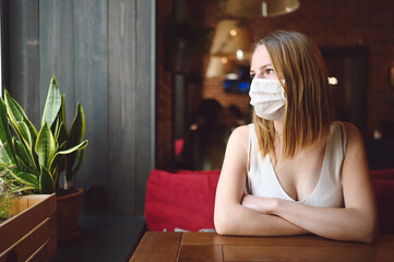 Young beautiful girl is sitting in a cafe or restaurant and waiting for her order wearing face protective mask for flu virus prevention. Corona virus and social distancing concept