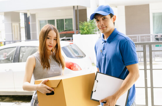 Smiling young Asian woman receiving package box from Asian delivery man in blue uniform at front of house and looking at camera.Delivery service , Onlines shopping concept.