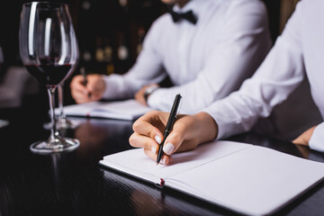 Cropped view of sommelier writing on notebook near colleague and glasses of wine on table