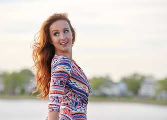 Portrait of stunning young woman with red hair standing in beautiful flowy print dress near small lake or bay at shore community