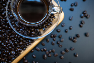 Top view part of black coffee in transparent cup and saucer with coffee beans on wooden tray background. Focus at the edge of glass for background blur.