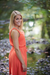 Fototapeta premium Beautiful young blonde woman poses outside in coral colored sundress stands along small stream in woods