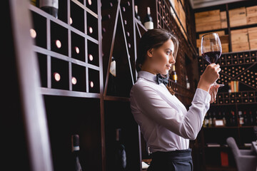 Selective focus of sommelier looking at wine in glass near racks with bottles