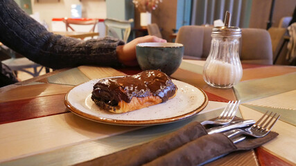 Dessert and cappuccino are on the table, male hands in the frame hold a cup. Chocolate baking with poppy seeds lies on a saucer in the foreground. Breakfast in a cafe, croissant and drink are shown.
