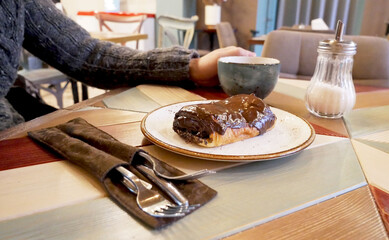 Dessert and cappuccino are on the table, male hands in the frame hold a cup. Chocolate baking with poppy seeds lies on a saucer in the foreground. Breakfast in a cafe, croissant and drink are shown.