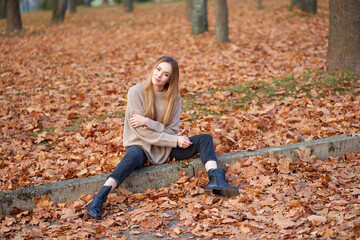 Pretty young blonde smiling woman wearing stylish oversize sweater, trendy black mom jeans and black leather boots posing in autumn park.