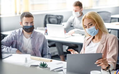 Meeting and discussing project during coronavirus epidemic. Young man and woman in protective masks look at laptop and communicate through glass board at workplace