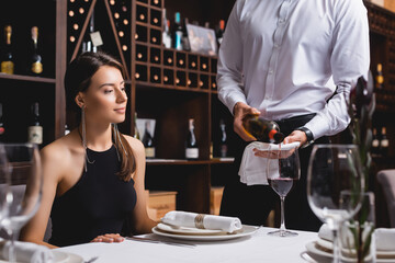 Selective focus of sommelier pouring wine in glass near young woman in restaurant