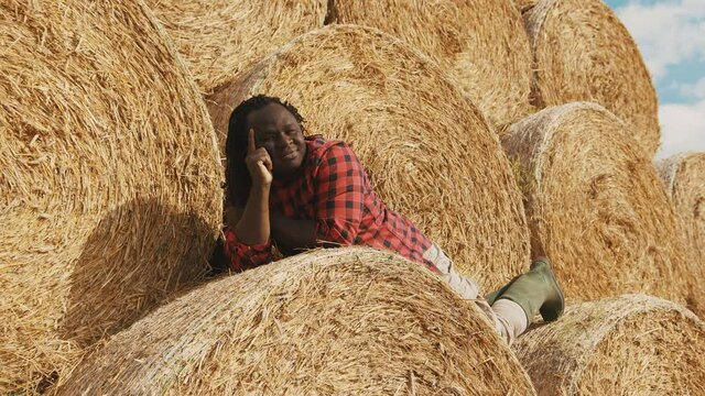 Young happy african farmer lying on the stack of hay rolls and resting. High quality 4k footage