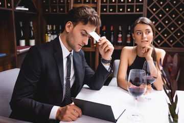 Selective focus of pensive man in suit holding credit card while looking at menu near glasses of wine and girlfriend in restaurant