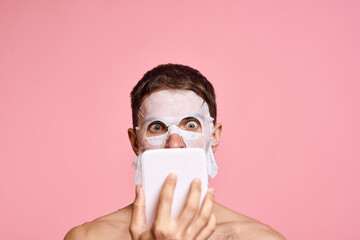 a man with a cleansing mask on his face examines himself in a mirror on a pink background