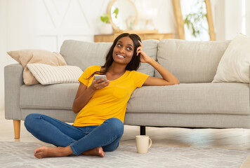 Obraz premium Beautiful black woman with smartphone sitting on floor in living room and studying online from home, copy space