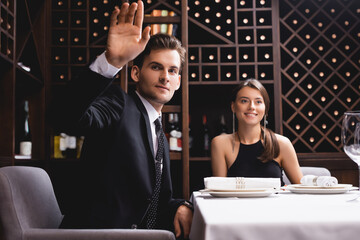 Selective focus of man in suit waving hand near elegant woman in restaurant