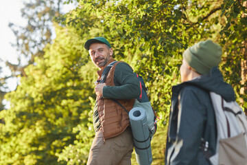 Naklejka na ściany i meble Portrait of happy father and son hiking together and talking while walking in forest with backpack, copy space
