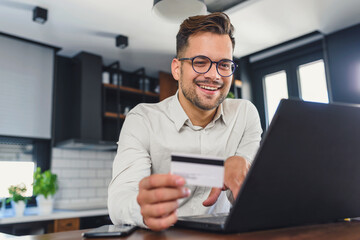 Young man holding credit card sitting in front of laptop computer at home, paying for online order. People, lifestyle, modern technologies and e-commerce concept. Online banking and shopping using mod