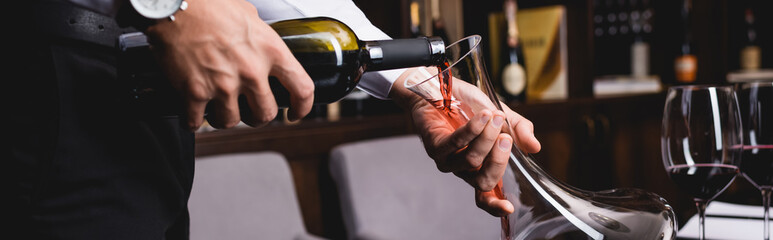 Panoramic shot of sommelier pouring wine in decanter near glasses in restaurant - obrazy, fototapety, plakaty