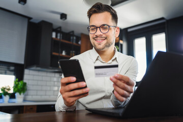 Young man holding credit card sitting in front of laptop computer at home, paying for online order. People, lifestyle, modern technologies and e-commerce concept. Online banking and shopping using mod