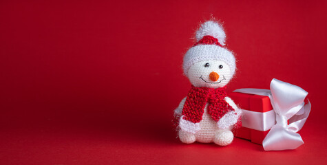 A snowman toy in a Santa Claus hat on a red background next to a red gift box tied with a white ribbon: space for text
