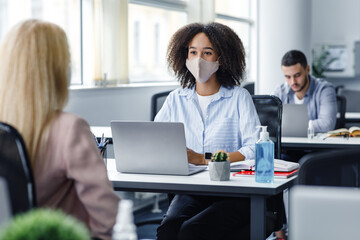 Work with client after quarantine and social distancing. Friendly african american woman in protective mask talking to european lady, sitting at workplace with laptop