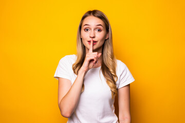 Portrait of a smiling young woman showing silence gesture and looking at camera isolated over yellow background