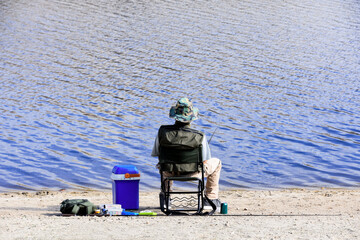Back foreground of a sitting man angling on the shore of a lake with his spare rod and tackle
