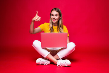 Happy young woman have idea sitting on the floor with crossed legs and using laptop on red background.