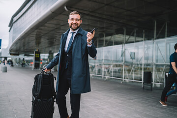 Half length portrait of cheerful businessman with baggage suitcase smiling at camera while waiting...