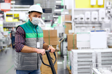 young caucasian warehouse worker in medical mask with hand pallet truck with package shipment...